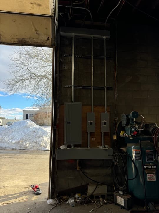 Commercial garage workshop with a freshly mounted electrical sub-panel and disconnects on a wood backboard, viewed through an open bay door onto a snowy parking lot.