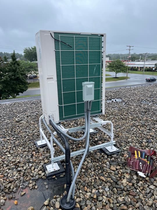 Large green commercial HVAC condenser unit on a metal stand on a gravel pad, with electrical disconnect frame and tools nearby.