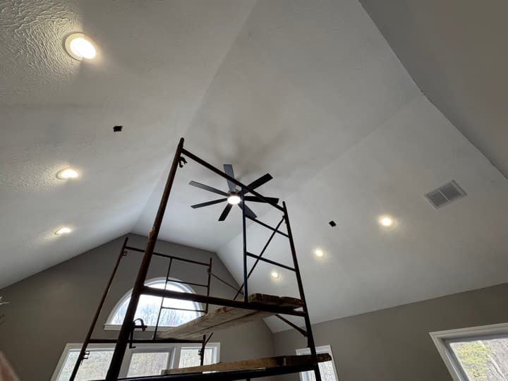Vaulted living-room ceiling with multiple recessed cans and a black center ceiling fan installed during finish work, with a scaffold tower in the foreground for ceiling access.