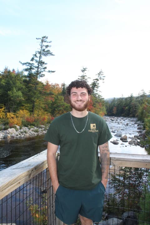 Brian Arlin standing on a wooden bridge over a New Hampshire river in early autumn, fall foliage and the White Mountains visible behind him.