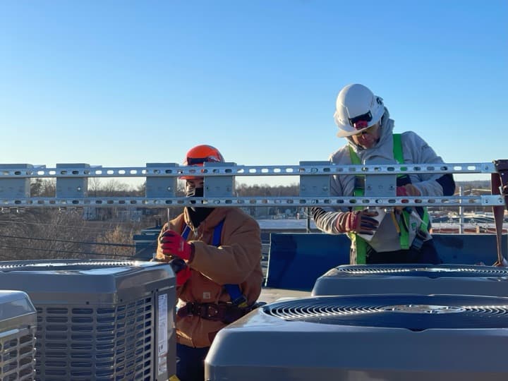 Brian on a commercial flat roof in his electrician PPE — orange helmet, fall-arrest harness, Carhartt jacket — installing rooftop HVAC support strut alongside a coworker.